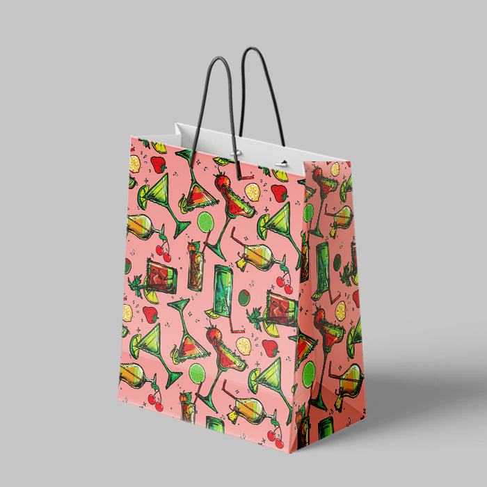 A pink paper shopping bag with a colorful cocktail-themed pattern, featuring black twisted paper handles, set against a solid gray background.