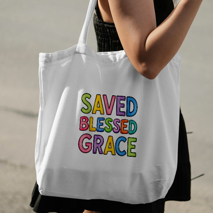 A white canvas tote bag with colorful bubble-letter text reading 'SAVED BLESSED GRACE' being carried on a person's shoulder against a blurred outdoor background.
