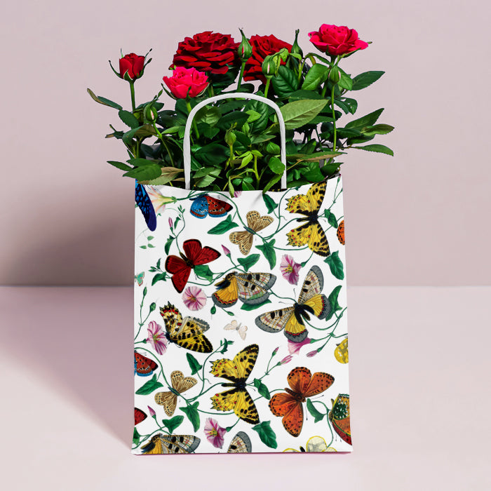 A white paper gift bag decorated with a colorful pattern of butterflies and small flowers, filled with red roses and green foliage, standing on a light pink surface against a pale pink background.