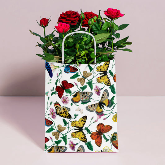 A white paper gift bag decorated with a colorful pattern of butterflies and small flowers, filled with red roses and green foliage, standing on a light pink surface against a pale pink background.
