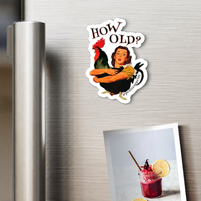 A die-cut magnet featuring a retro-style illustration of a woman hugging a rooster with the text 'HOW OLD?' above and 'permanently' below, shown attached to a stainless steel refrigerator door next to a photo print.