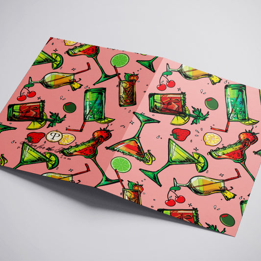 A folded greeting card featuring a vibrant pink background with a repeating pattern of colorful cocktail illustrations, displayed on a neutral light gray surface.
