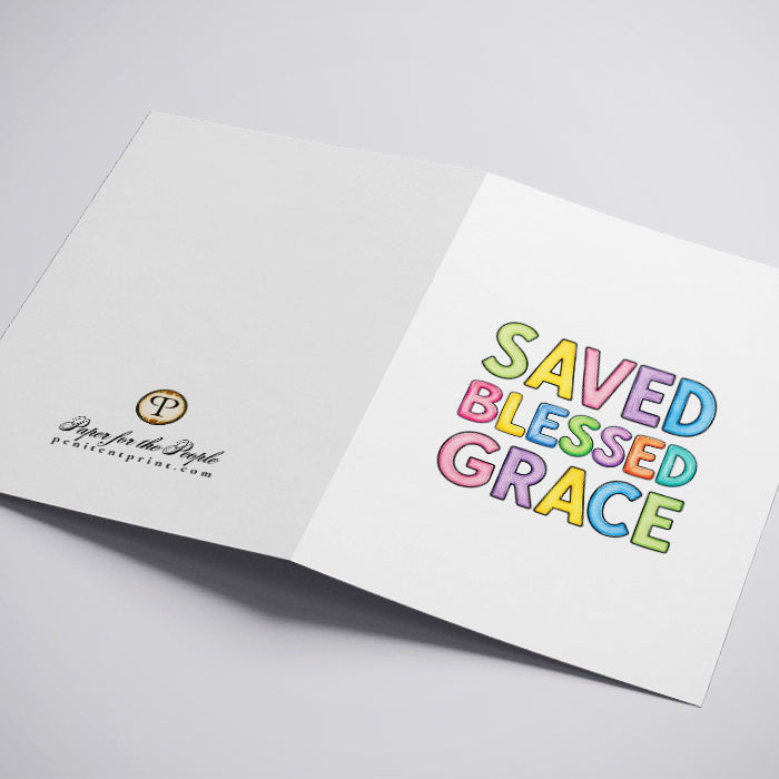 An open white greeting card on a light gray background. The left side shows the inside with a small circular logo and the text 'Saved Blessed Grace'