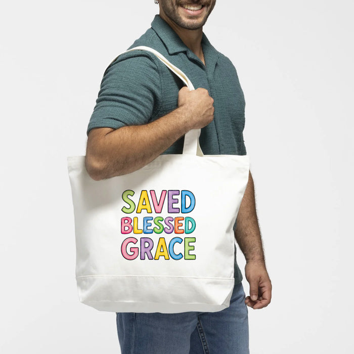 A man wearing a green polo shirt and blue jeans carries a white canvas tote bag over his shoulder. The bag features the text 'SAVED BLESSED GRACE' printed in a colorful, rainbow-like outline font.