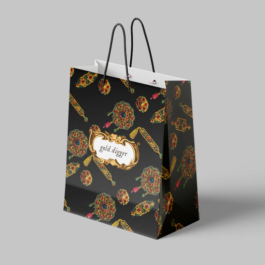 Black paper giftbag with gold and multicolor medieval jewel pattern and 'gold digger' text on a gray background