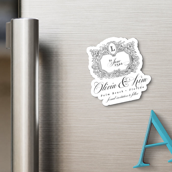 A custom die-cut white refrigerator magnet with black script and ornate border details, displayed on a brushed stainless steel refrigerator door next to a blue letter prop.