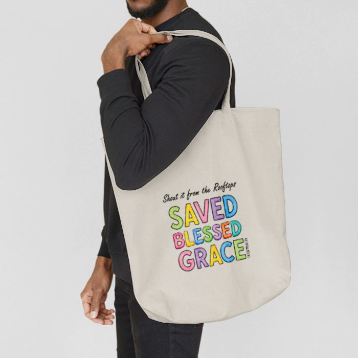 A man wearing a black long sleeve t-shirt and black jeans carries a white canvas tote bag over his shoulder. The bag features the text 'Shout it from the Rooftops! SAVED BLESSED GRACE' in colorful pastel letters, printed in a rainbow-like outline font.